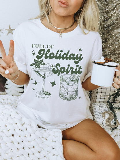 Oversized white crew neck tshirt worn as a dress. This holiday graphic tee features a minimalist tall cocktail glass as well as a short whiskey glass with the words full of holiday spirit in green.