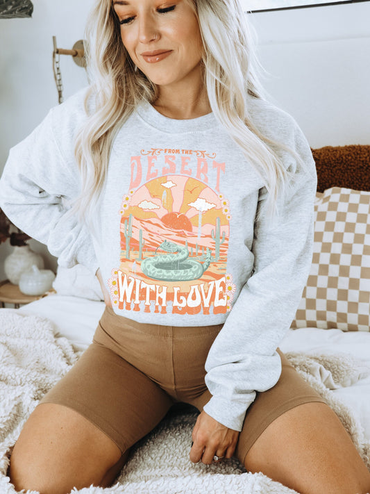 Boho bedroom with neutral aesthetic featuring a model wearing an oversized, long sleeve crew neck pullover. This trendy sweatshirt has a distressed desert image featuring a snake, cactus, mountains and a setting sun in earthy tones.