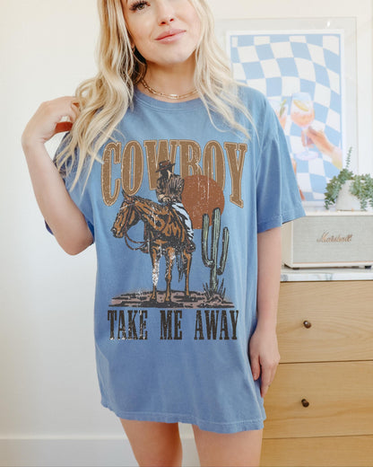 Model is in an oversized blue crewneck t-shirt featuring a rustic country western graphic in neutral tones. Graphic contains a cowboy at sunset in the desert with the words, cowboy take me away.