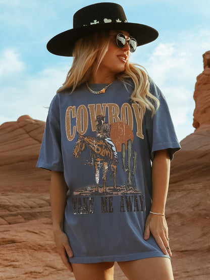 Model is in an oversized denim blue crewneck t-shirt featuring a rustic country western graphic in neutral tones. Graphic contains a cowboy at sunset in the desert with the words, cowboy take me away.