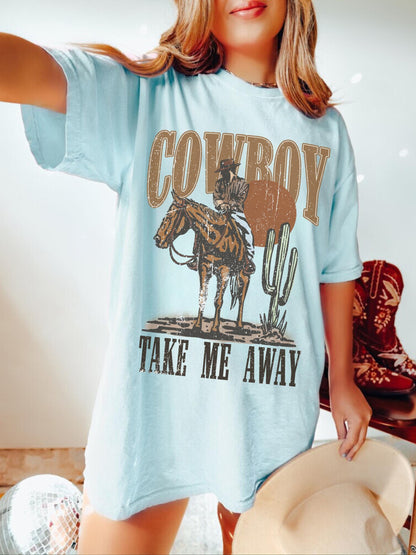 Model is in an oversized light blue crewneck t-shirt featuring a rustic country western graphic in neutral tones. Graphic contains a cowboy at sunset in the desert with the words, cowboy take me away.