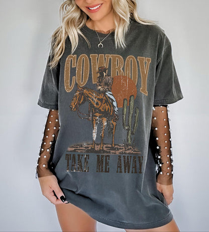 Model is in an oversized dark grey distressed crewneck t-shirt featuring a rustic country western graphic in neutral tones. Graphic contains a cowboy at sunset in the desert with the words, cowboy take me away.