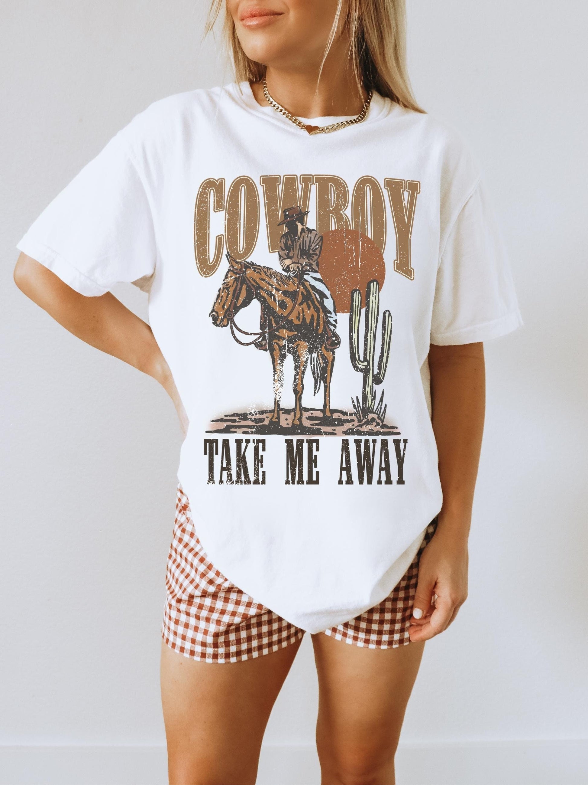 Model is in an oversized white crewneck t-shirt featuring a rustic country western graphic in neutral tones. Graphic contains a cowboy at sunset in the desert with the words, cowboy take me away.